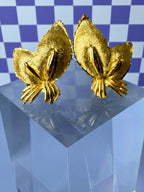 Pair of gold leaf-shaped earrings on a clear stand with a blue checkered background