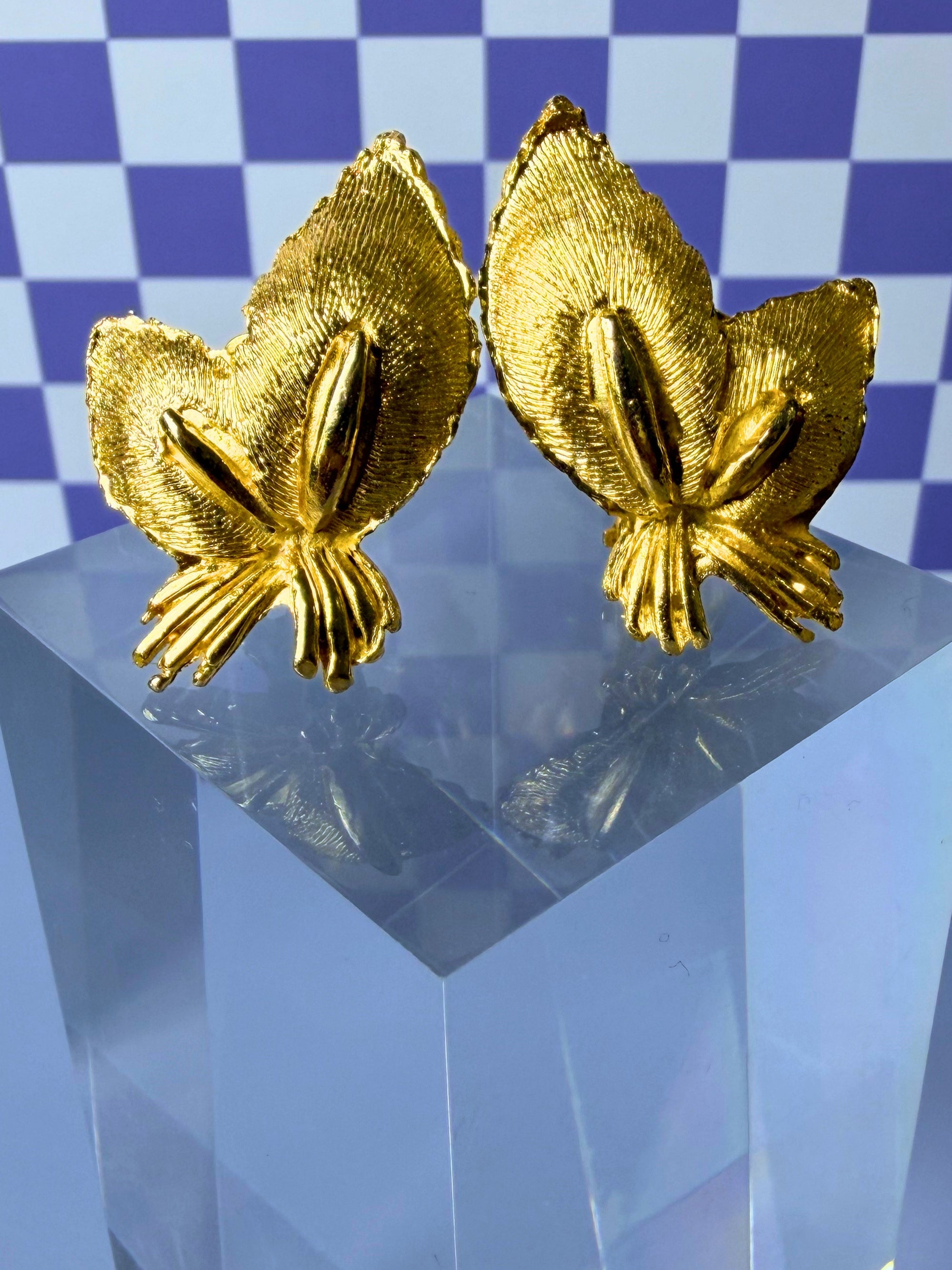 Pair of gold leaf-shaped earrings on a clear stand with a blue checkered background