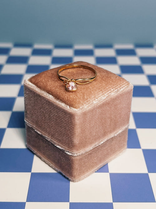 Gold ring with a pink gemstone on a pink velvet cushion against a blue and white checkered background