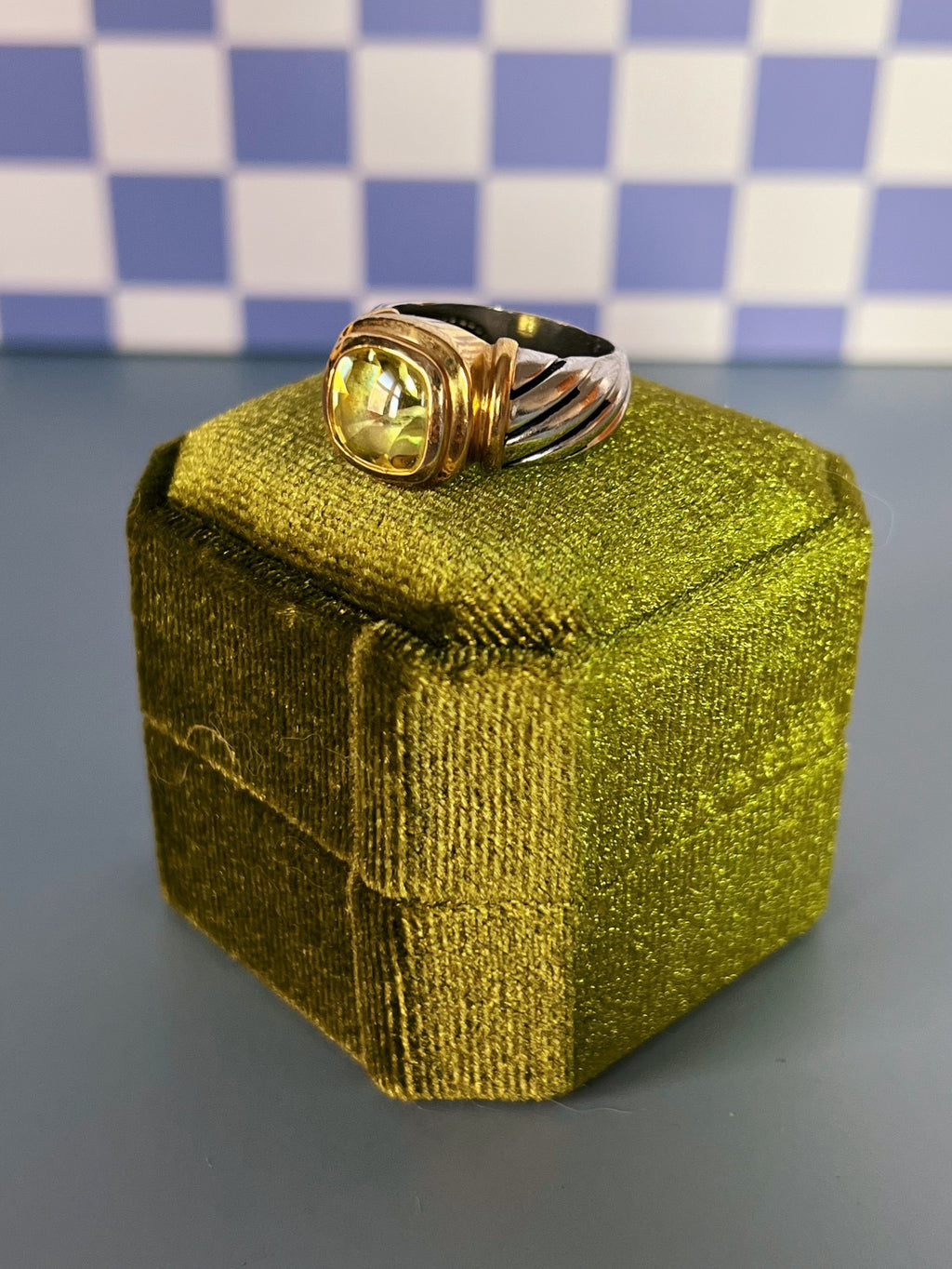 Gold ring with a green crystal on a green box against a checkered background