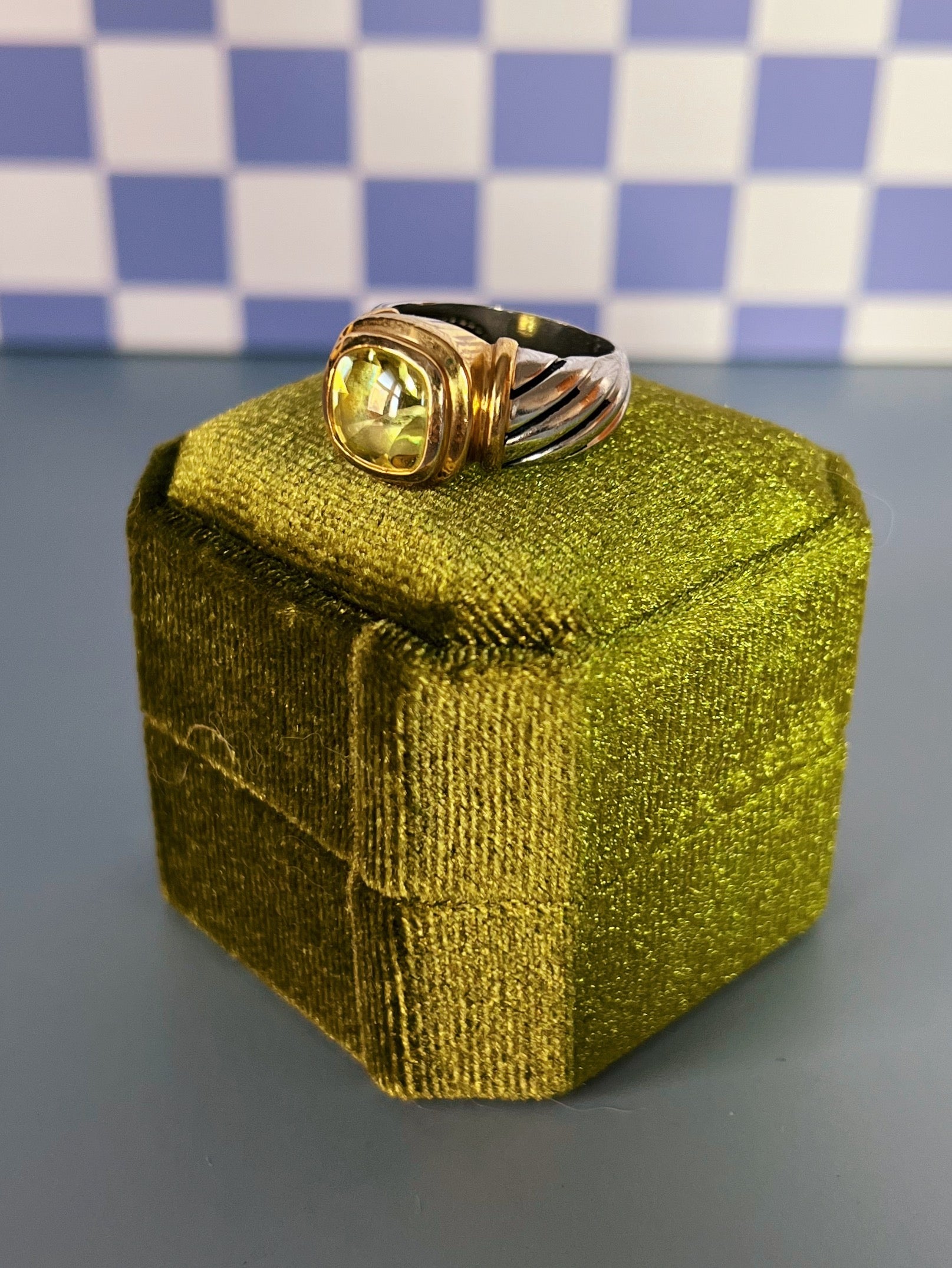 Gold ring with a green crystal on a green box against a checkered background